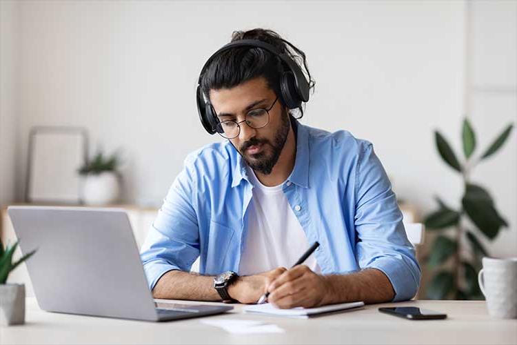 图为一名黑发青年, 一个胡子, 和眼镜, 在打开的笔记本电脑前做笔记.